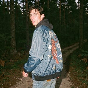 May include: A blue bomber jacket with a tiger design on the back. The jacket has a black collar and cuffs. The person in the image is wearing a gray hoodie underneath the jacket and blue jeans.