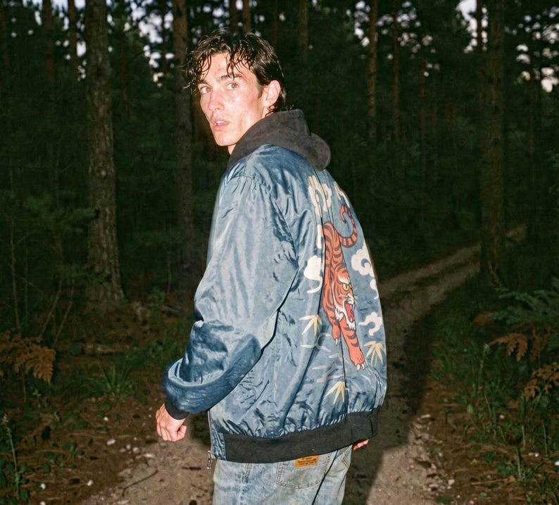May include: A blue bomber jacket with a tiger design on the back. The jacket has a black collar and cuffs. The person in the image is wearing a gray hoodie underneath the jacket and blue jeans.