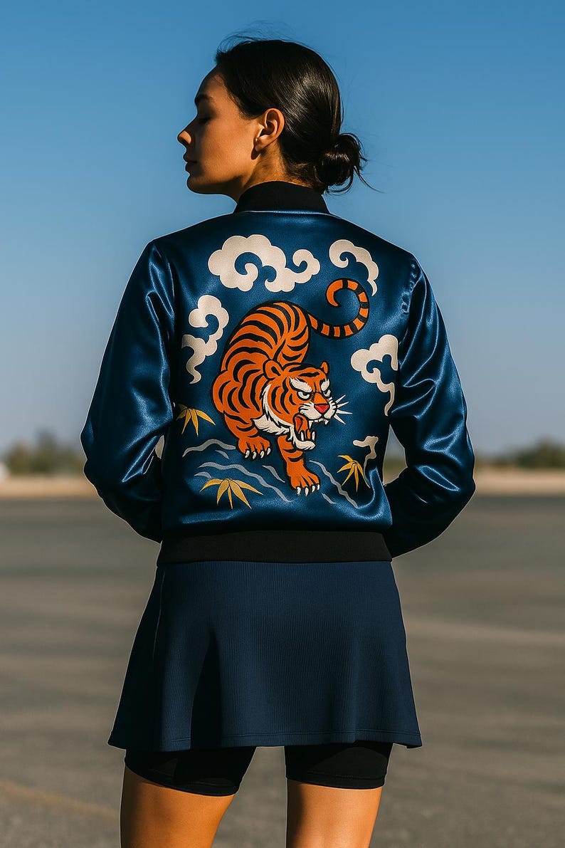 May include: A navy blue satin bomber jacket with a tiger design on the back. The tiger is orange and black, surrounded by white clouds. The jacket has a black collar and cuffs. The model is wearing a navy blue skirt.