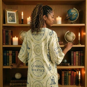 May include: A cream-colored short-sleeved shirt featuring a repeating celestial print with blue and gold circular diagrams, star charts, and the text "COSMOS SOLSTICE" and "AURA" printed throughout the fabric, worn by a person facing a bookshelf.