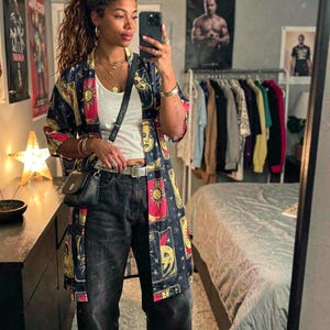 May include: A navy blue kimono-style jacket with a celestial design featuring suns, moons, and faces. The jacket is paired with a white top, black jeans, and a small black purse. The outfit is accessorized with jewelry.