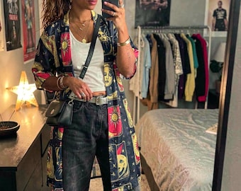 May include: A navy blue kimono-style jacket with a celestial design featuring suns, moons, and faces. The jacket is paired with a white top, black jeans, and a small black purse. The outfit is accessorized with jewelry.