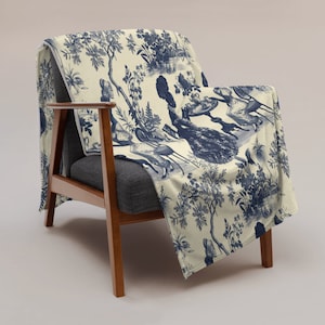 Op de afbeelding: Een blauwe en witte deken met een toile-patroon met figuren in een tuinsetting. De deken is over een houten fauteuil gelegd.