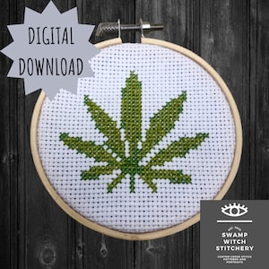 May include: A green marijuana leaf cross stitch pattern in a wooden embroidery hoop. The pattern is on white Aida cloth. The hoop is hanging on a dark wood background. The text "Swamp Witch Stitchery" is visible in the bottom right corner of the image.