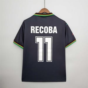 May include: Dark navy football shirt with the name "RECOBA" and the number "11" in white. The collar and sleeve cuffs have green, yellow, and red trim. The shirt is displayed on a wooden hanger.