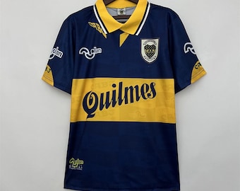 May include: A navy blue and yellow soccer jersey with the word "Quilmes" across the chest. The jersey has a yellow collar and sleeves with white trim. The Boca Juniors team crest is on the chest.