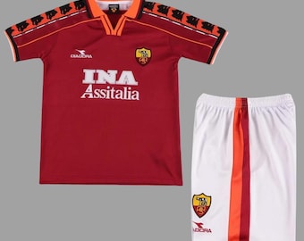 May include: A red and orange soccer jersey and white shorts set. The jersey has the text "INA Assitalia" and a team crest. The shorts have orange and red stripes down the side. The set is made by Diadora.