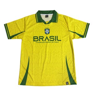 Puede incluir: Camiseta de fútbol amarilla con detalles verdes, con la palabra "BRASIL" en letras verdes. La camiseta presenta el logo de la Confederación Brasileña de Fútbol y un patrón de diamantes. Manga corta, con cuello.