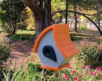 May include: A gray and orange birdhouse with a black circular entrance hangs from a black metal bracket. The birdhouse has an orange roof and is surrounded by green plants and colorful flowers in a garden setting.