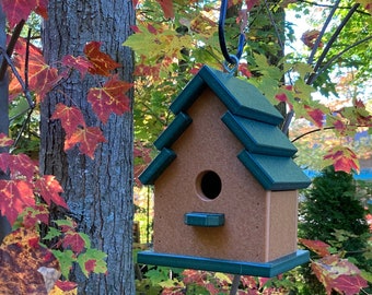 May include: A brown and green birdhouse with a green roof hangs from a tree branch. The tree has red, yellow, and green leaves.