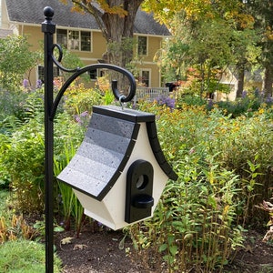 May include: A white and black birdhouse with a black roof hangs from a black metal stand in a garden setting. The birdhouse has a small opening for birds to enter.
