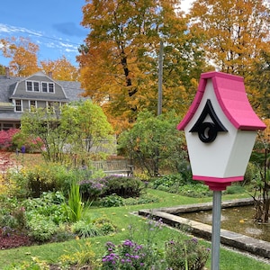 May include: A pink and white birdhouse with a black roof and a black circle on the front. The birdhouse is mounted on a metal pole in a garden setting with a pond and green grass.