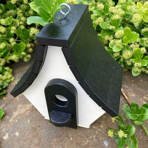 May include: A black and white birdhouse with a peaked roof and a small entrance hole. The birdhouse has a metal hook for hanging.