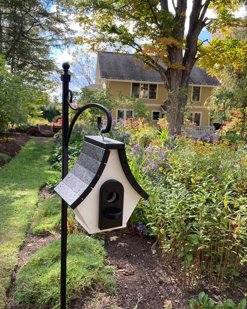 May include: A black and white birdhouse with a shingled roof sits on a black metal pole in a garden setting. The birdhouse has a small opening for birds to enter.