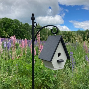Op de afbeelding: Een zwarte metalen vogelhuisje met een grijs dak hangt aan een zwarte metalen paal. Het vogelhuisje is omringd door een veld met paarse en roze lupine bloemen.