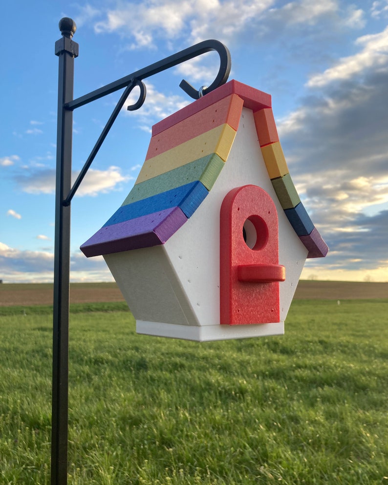 May include: A rainbow-colored birdhouse with a red door and a white base. The birdhouse is hanging from a black metal hook.