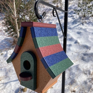 Op de afbeelding: Een vogelhuisje met een veelkleurig dak, met tinten rood, blauw en groen. Het vogelhuisje is gemaakt van hout en heeft een kleine opening waar vogels naar binnen kunnen. Het hangt aan een zwarte metalen haak.
