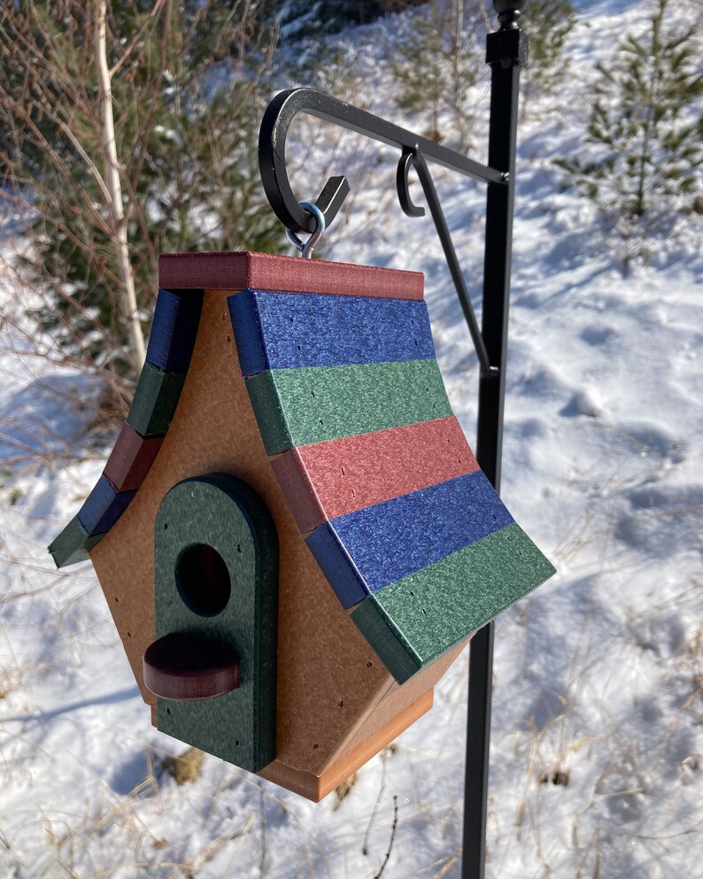 Op de afbeelding: Een vogelhuisje met een veelkleurig dak, met tinten rood, blauw en groen. Het vogelhuisje is gemaakt van hout en heeft een kleine opening waar vogels naar binnen kunnen. Het hangt aan een zwarte metalen haak.
