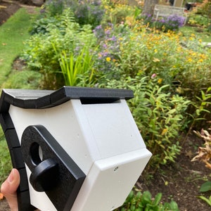May include: A white birdhouse with a black roof and trim. The birdhouse has a round opening for birds to enter. The birdhouse is held in a person's hand.