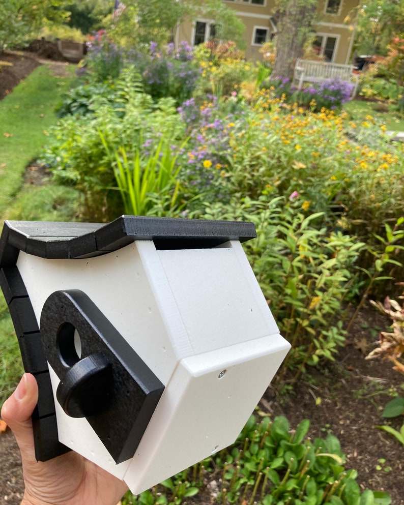 May include: A white birdhouse with a black roof and trim. The birdhouse has a round opening for birds to enter. The birdhouse is held in a person's hand.