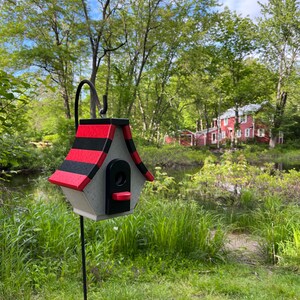 Op de afbeelding: Een zwart-rood gestreept vogelhuisje met een grijze basis en een zwarte opening. Het vogelhuisje is gemonteerd op een zwarte metalen paal en staat voor een groene graszone.