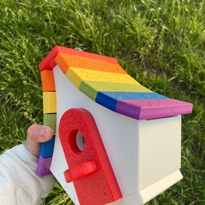 May include: A rainbow-colored birdhouse with a red door. The birdhouse is made of wood and has a white base. The roof is made of colorful wooden shingles.
