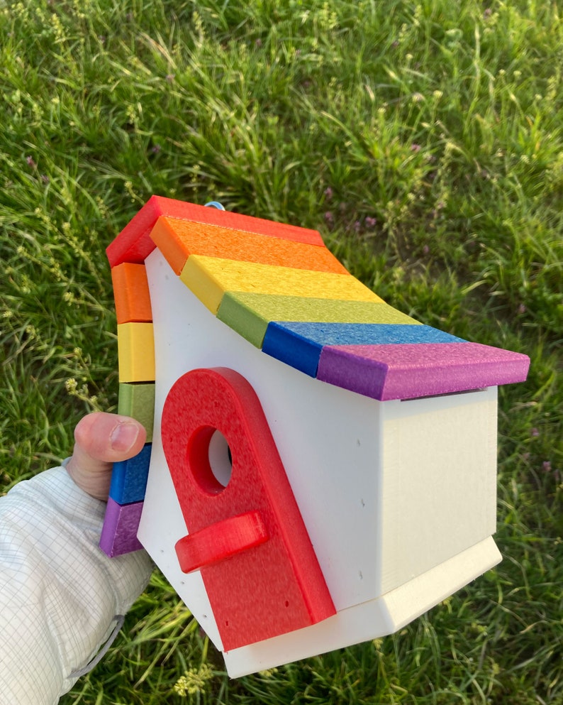 May include: A rainbow-colored birdhouse with a red door. The birdhouse is made of wood and has a white base. The roof is made of colorful wooden shingles.