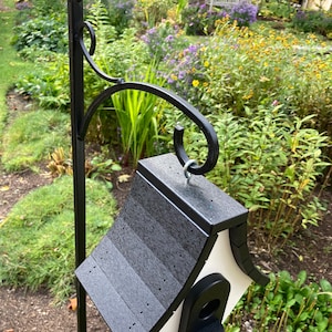 May include: A black and white birdhouse with a black shingled roof hangs from a black metal hook. The birdhouse is mounted on a black metal pole.