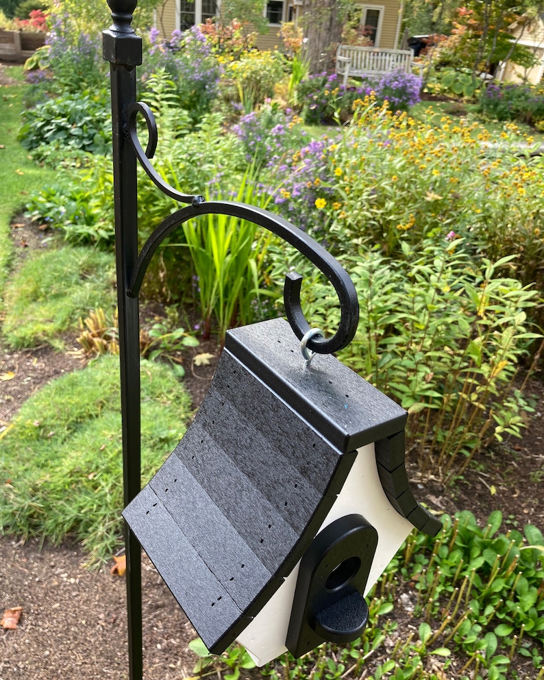 May include: A black and white birdhouse with a black shingled roof hangs from a black metal hook. The birdhouse is mounted on a black metal pole.