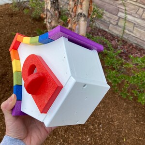 May include: A white birdhouse with a rainbow-colored roof. The roof is made of individual pieces of foam in different colors, including red, orange, yellow, green, blue, indigo, and violet. The birdhouse has a small, red, round entrance hole.