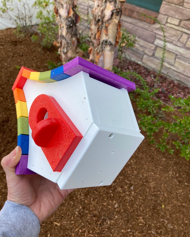 May include: A white birdhouse with a rainbow-colored roof. The roof is made of individual pieces of foam in different colors, including red, orange, yellow, green, blue, indigo, and violet. The birdhouse has a small, red, round entrance hole.