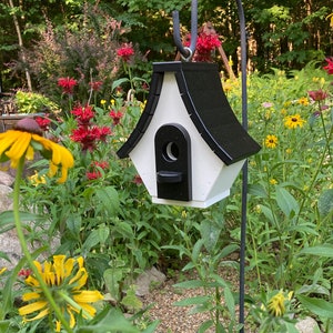 May include: A white and black birdhouse with a black roof and a black entrance hole. The birdhouse is hanging from a black metal hook in a garden setting.