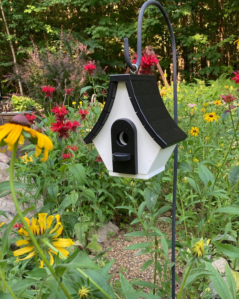 May include: A white and black birdhouse with a black roof and a black entrance hole. The birdhouse is hanging from a black metal hook in a garden setting.