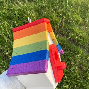 May include: A rainbow-colored birdhouse with a red entrance hole. The birdhouse is made of wood and has a white base. It is decorated with a rainbow pattern and has a metal ring on top for hanging.