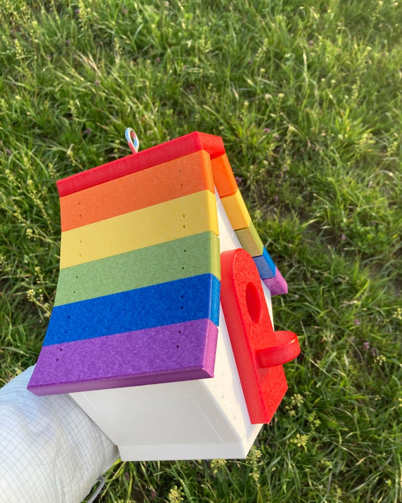 May include: A rainbow-colored birdhouse with a red entrance hole. The birdhouse is made of wood and has a white base. It is decorated with a rainbow pattern and has a metal ring on top for hanging.
