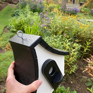 May include: A black and white birdhouse with a rounded roof and a small entrance hole. The birdhouse has a metal hook on top for hanging.