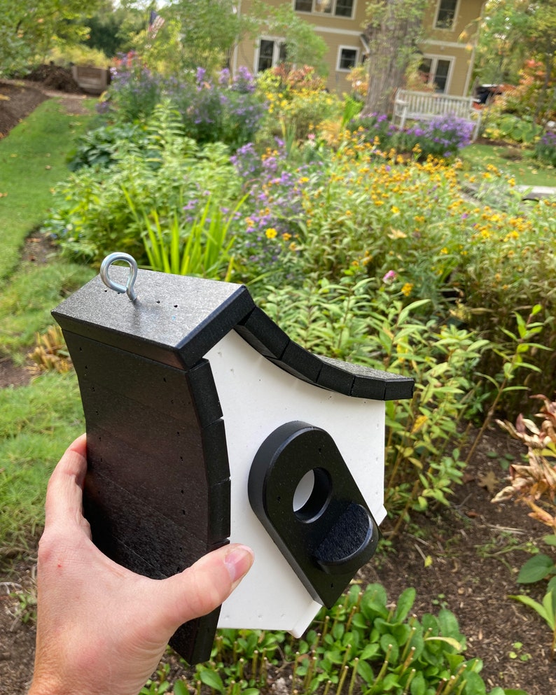 May include: A black and white birdhouse with a rounded roof and a small entrance hole. The birdhouse has a metal hook on top for hanging.