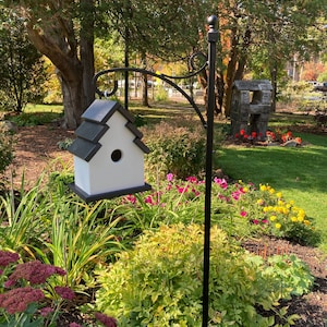 May include: A white birdhouse with a black roof sits on a black metal stand in a garden setting. The birdhouse is surrounded by green grass, flowers, and trees.