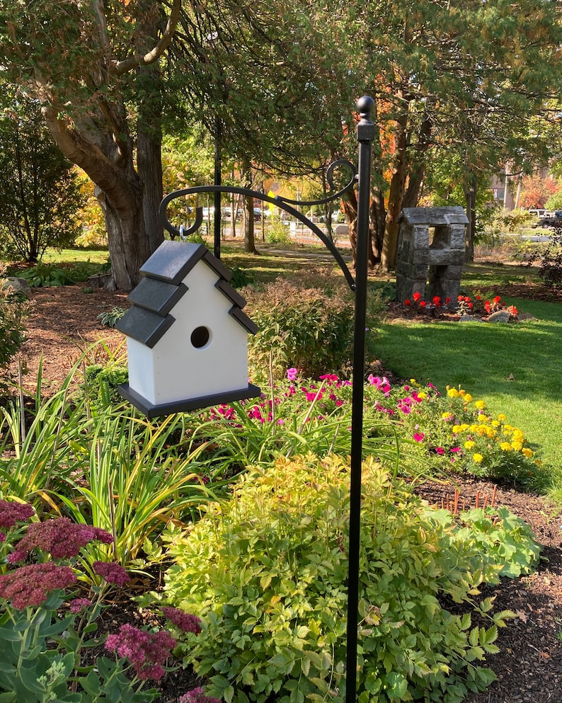 May include: A white birdhouse with a black roof sits on a black metal stand in a garden setting. The birdhouse is surrounded by green grass, flowers, and trees.