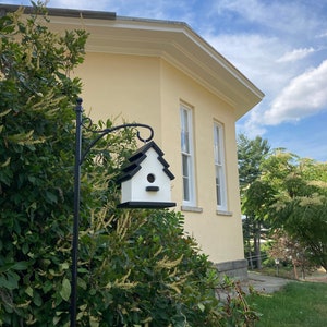 May include: A white birdhouse with a black roof and a small opening for birds to enter. The birdhouse is mounted on a black metal pole and is attached to a yellow building with white trim.
