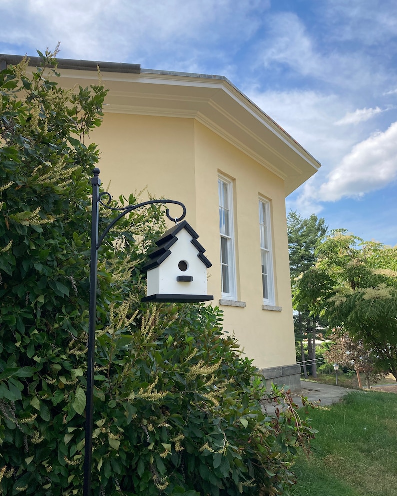May include: A white birdhouse with a black roof and a small opening for birds to enter. The birdhouse is mounted on a black metal pole and is attached to a yellow building with white trim.