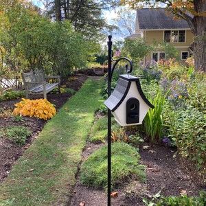 May include: A black metal birdhouse feeder with a white roof and a small opening for birds to enter. The feeder is mounted on a black metal pole and is surrounded by green grass and flowers.