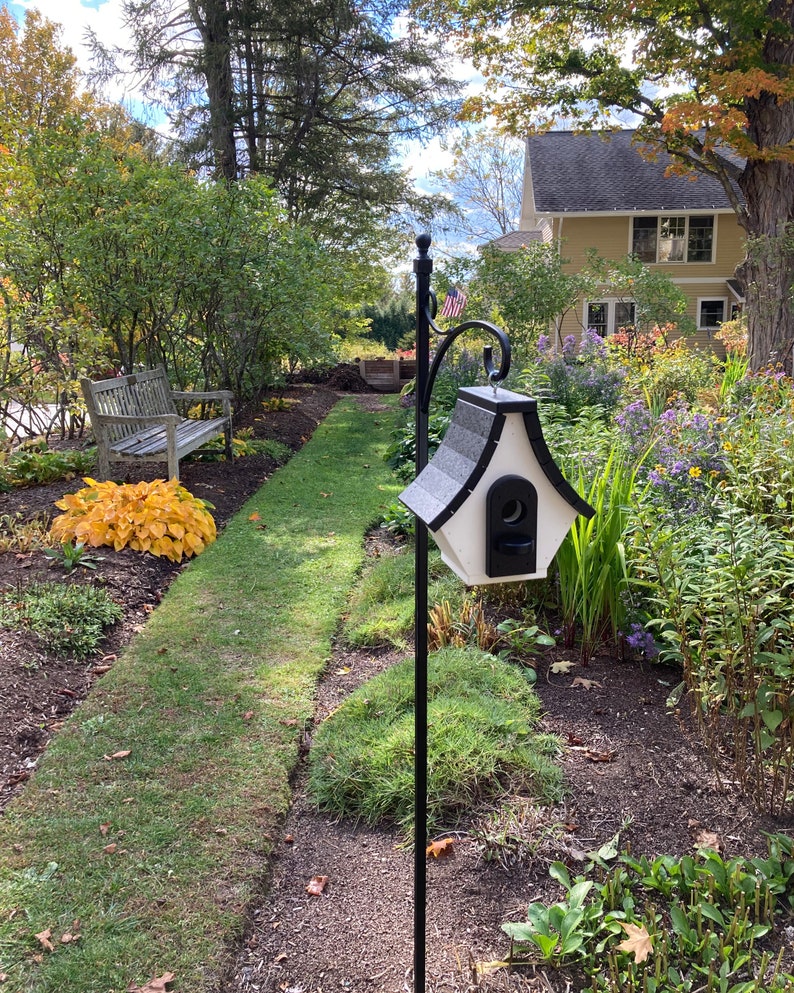 May include: A black metal birdhouse feeder with a white roof and a small opening for birds to enter. The feeder is mounted on a black metal pole and is surrounded by green grass and flowers.