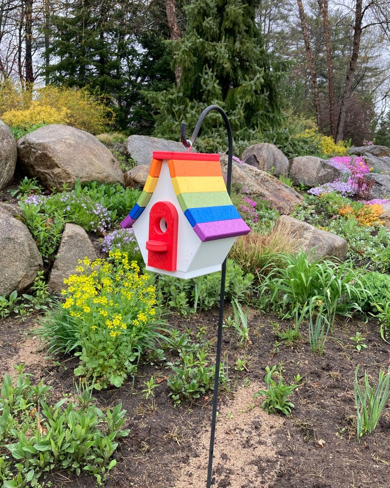 May include: A rainbow-colored birdhouse with a red door and a white base. The birdhouse is mounted on a black metal pole and is surrounded by green plants.