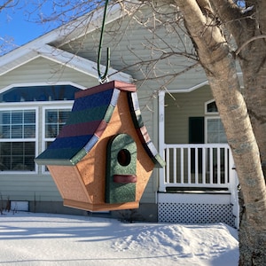 Op de afbeelding: Een houten vogelhuisje met een groen, blauw en rood dak. Het vogelhuisje hangt aan een tak en heeft een kleine opening waar vogels naar binnen kunnen.