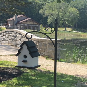 May include: A white birdhouse with a black roof hangs from a black metal stand. The birdhouse has a small opening for birds to enter. The birdhouse is in front of a stone bridge and a pond with green reeds.