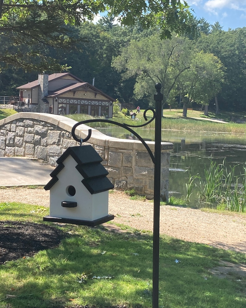 May include: A white birdhouse with a black roof hangs from a black metal stand. The birdhouse has a small opening for birds to enter. The birdhouse is in front of a stone bridge and a pond with green reeds.