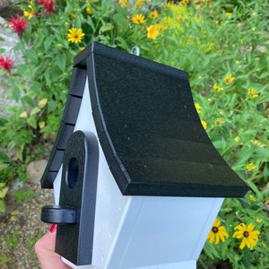 May include: A white birdhouse with a black roof and a black entrance hole. The birdhouse is made of wood and has a simple design.