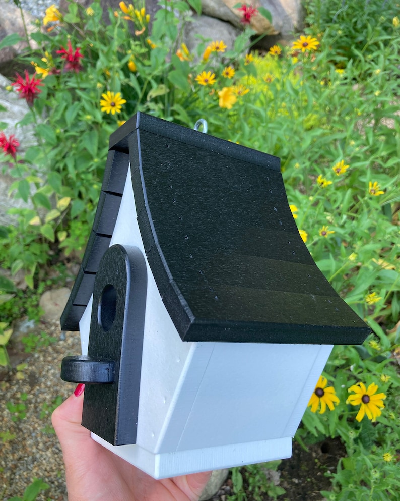 May include: A white birdhouse with a black roof and a black entrance hole. The birdhouse is made of wood and has a simple design.