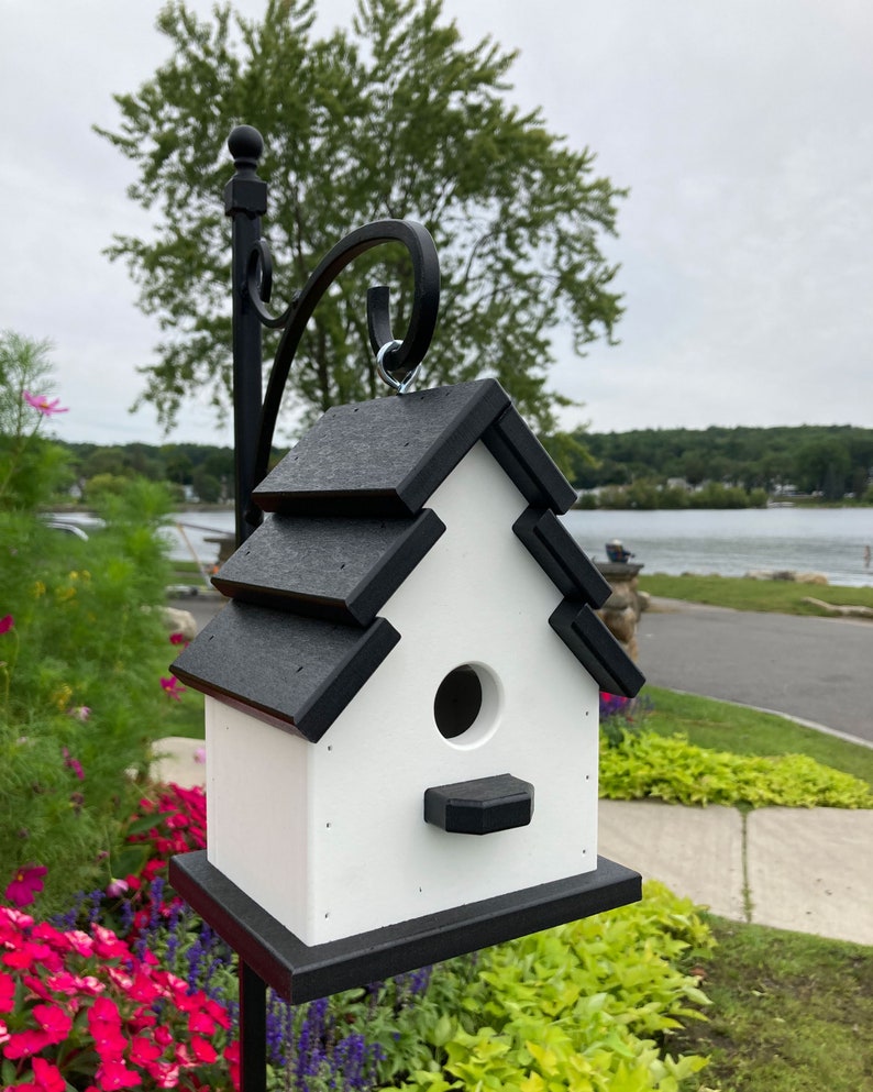 May include: A black and white birdhouse with a black roof and a white body. The birdhouse is hanging from a black metal hook. The birdhouse has a round entrance hole and a small landing platform.